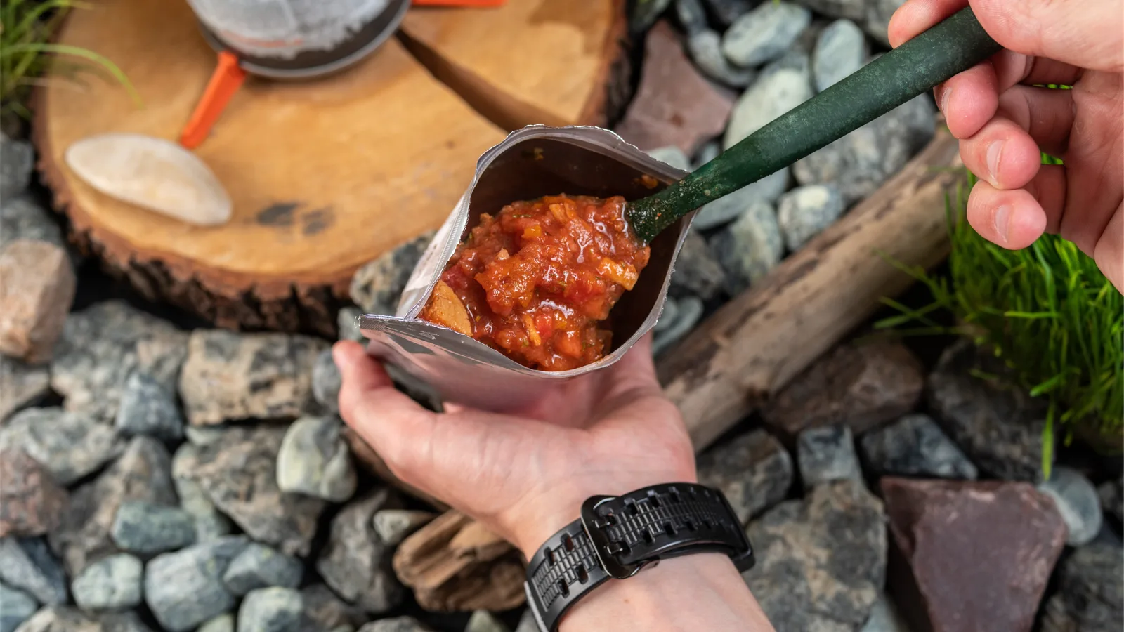 Hands holding an open silver pouch of rehydrated chilli beside a camp stove on a bed of river rock.